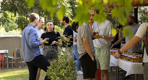42malaga_becalanoria Group of casually dressed adults chatting and holding drinks at an outdoor daytime party near a table of food.