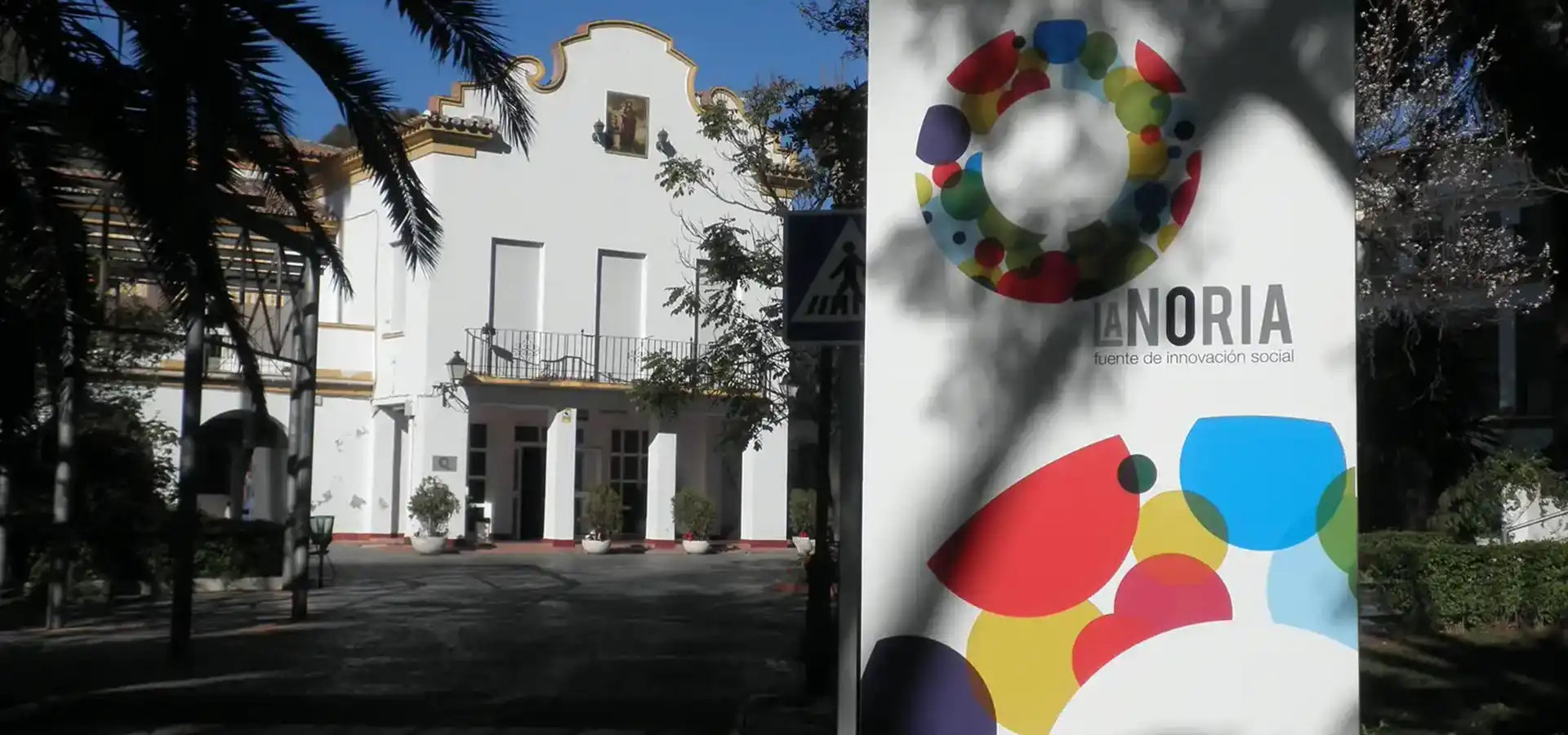 42malaga_lanoria White building with an arched top, palm trees in foreground, and a tall sign on the right displaying a colorful circular logo and the words 'ANORIA fuente de innovación social'