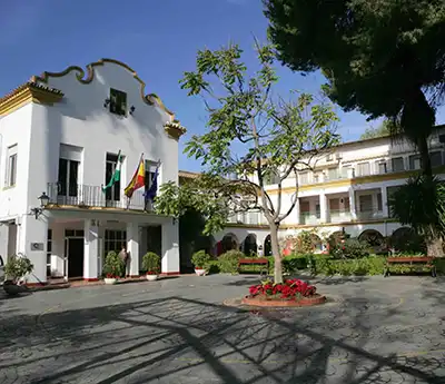 42malaga_solicitabecalanoria White colonial-style building with an arched porch, flags on the balcony, and a courtyard featuring a tree and a flower bed.